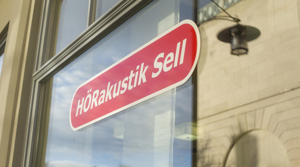 Schaufenster mit rotem Schild und weißer Aufschrift HÖRakustik Sell für Hörakustikgeschäft.